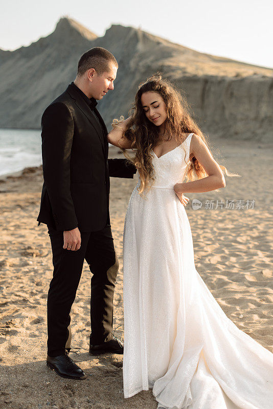 Attractive bride and groom pose on the beach. The bride in a luxurious dress stands on the sand among the mountains.